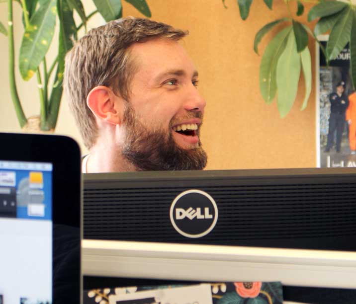 Digital designer, Ryan McLarty, sitting behind his computer, smiling and laughing while looking away from the camera. 